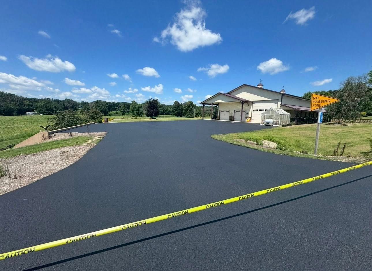 A newly paved, dark asphalt driveway leads to a suburban house under a blue, partly cloudy sky, with caution tape nearby.