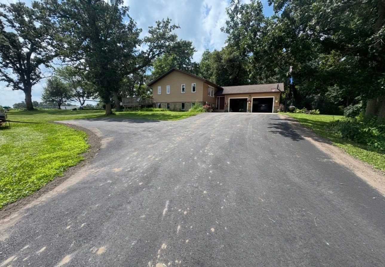 A tan house with a two-car garage sits at the end of a paved driveway, surrounded by green grass and large trees.