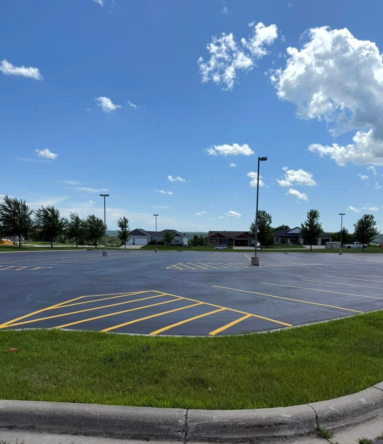Empty parking lot with yellow-painted lines on asphalt, surrounded by green grass and trees under a clear blue sky.