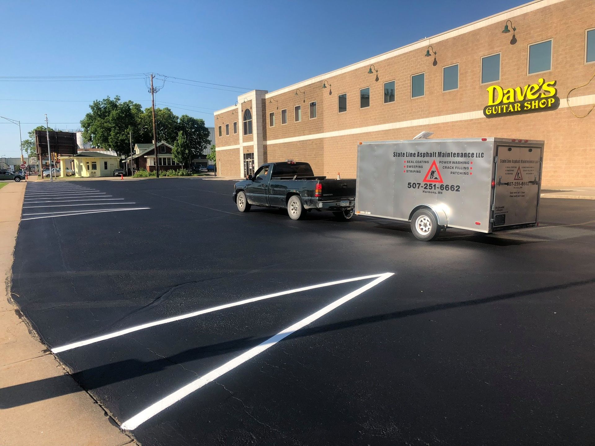 A truck towing a service trailer is parked on a freshly paved, marked parking lot in front of a brick building.