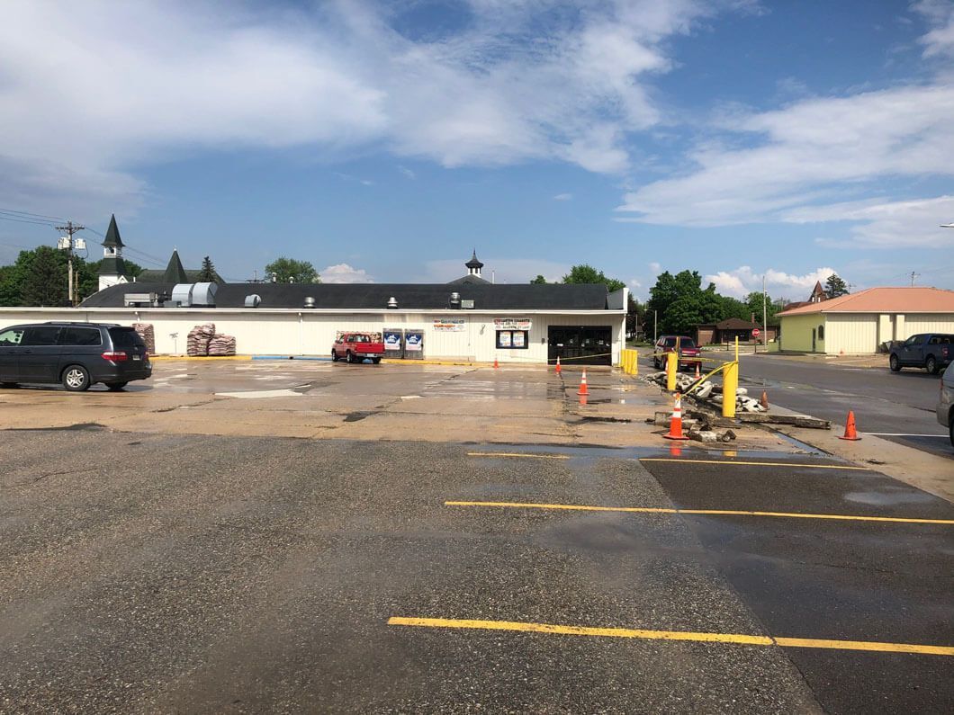 A paved, partially flooded parking lot in front of a one-story commercial building under a blue sky with scattered clouds.