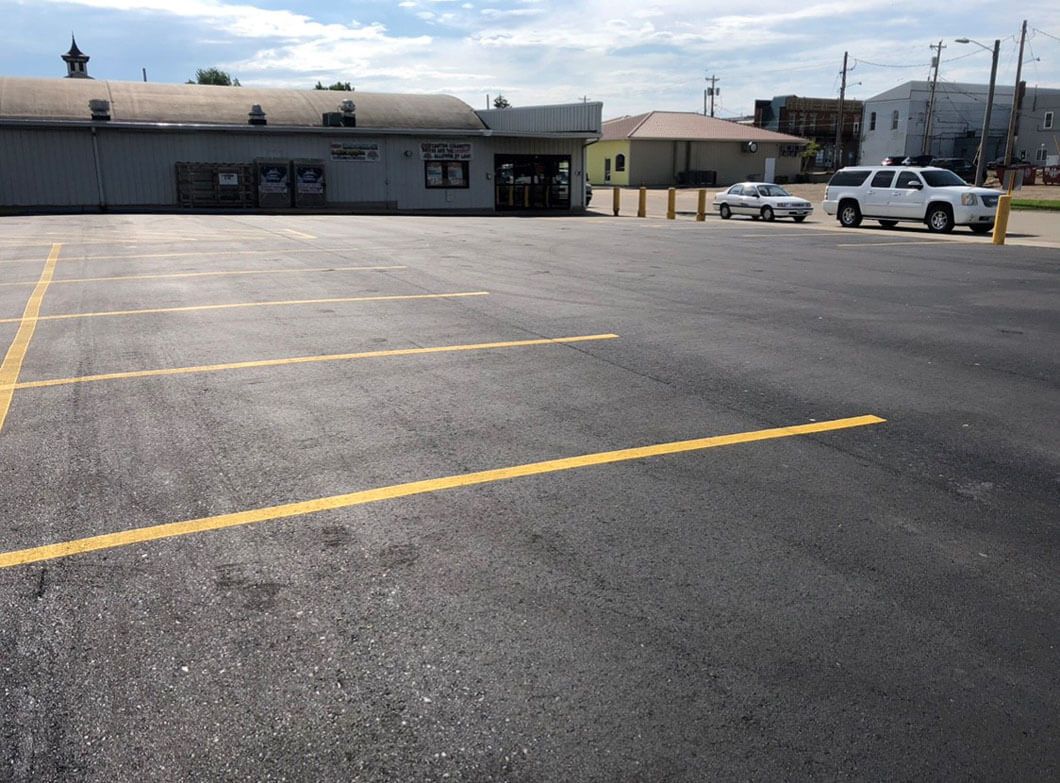 A paved parking lot with yellow painted lines, a grey building on the left, and a white SUV parked in the distance.