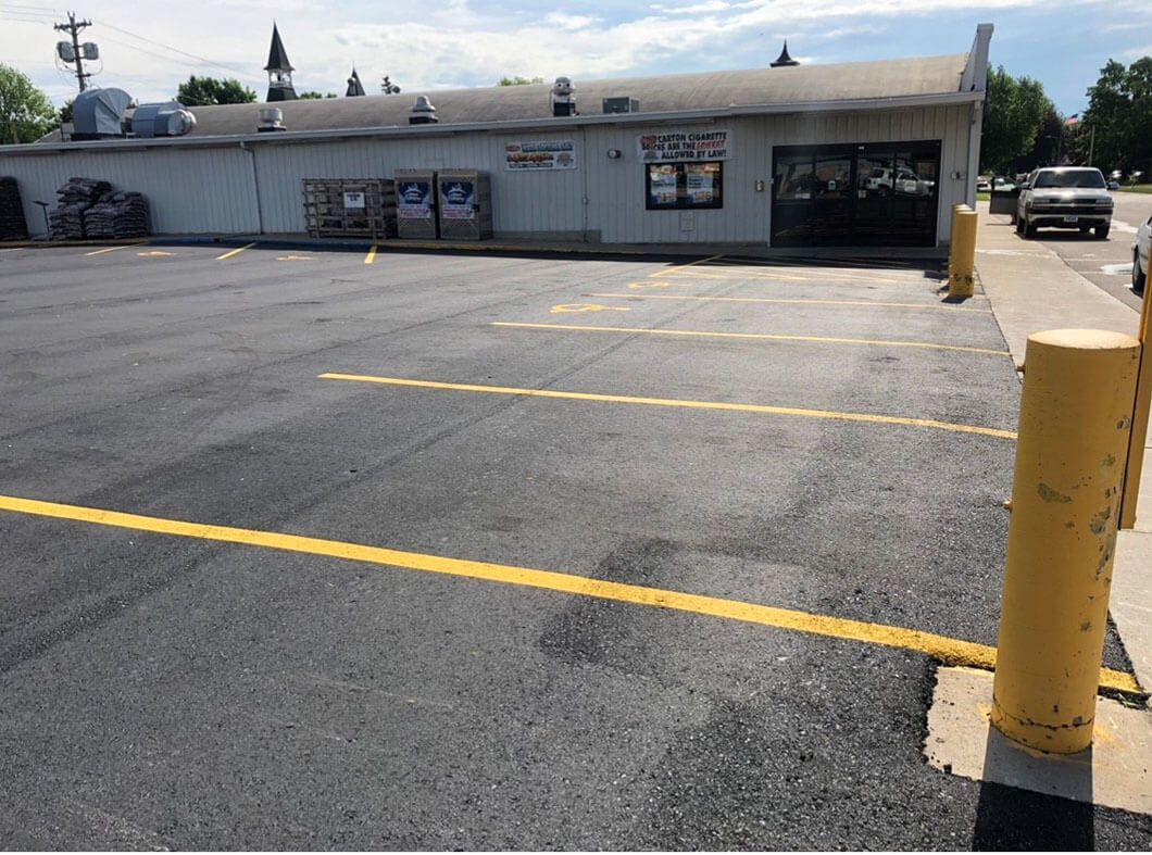 A paved parking lot with yellow-painted lines in front of a gray, single-story commercial building under a sunny sky.