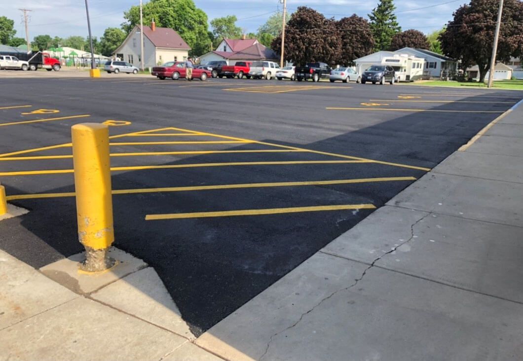 A freshly paved asphalt parking lot with yellow painted hash marks and bollards, next to a concrete sidewalk.