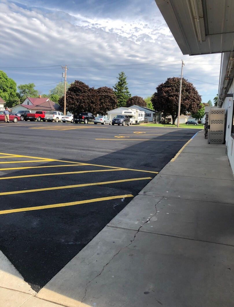 A view from a sidewalk overlooking a newly paved parking lot with yellow-painted safety markings in front of a building.