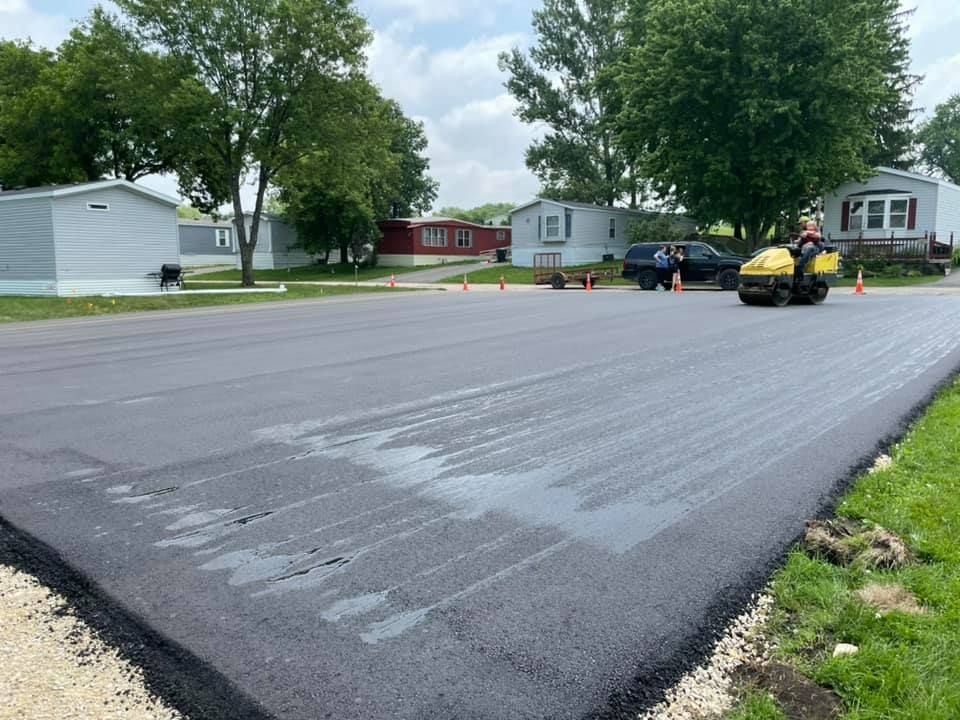 A bright yellow asphalt roller sits on a freshly paved black surface in a residential mobile home park.