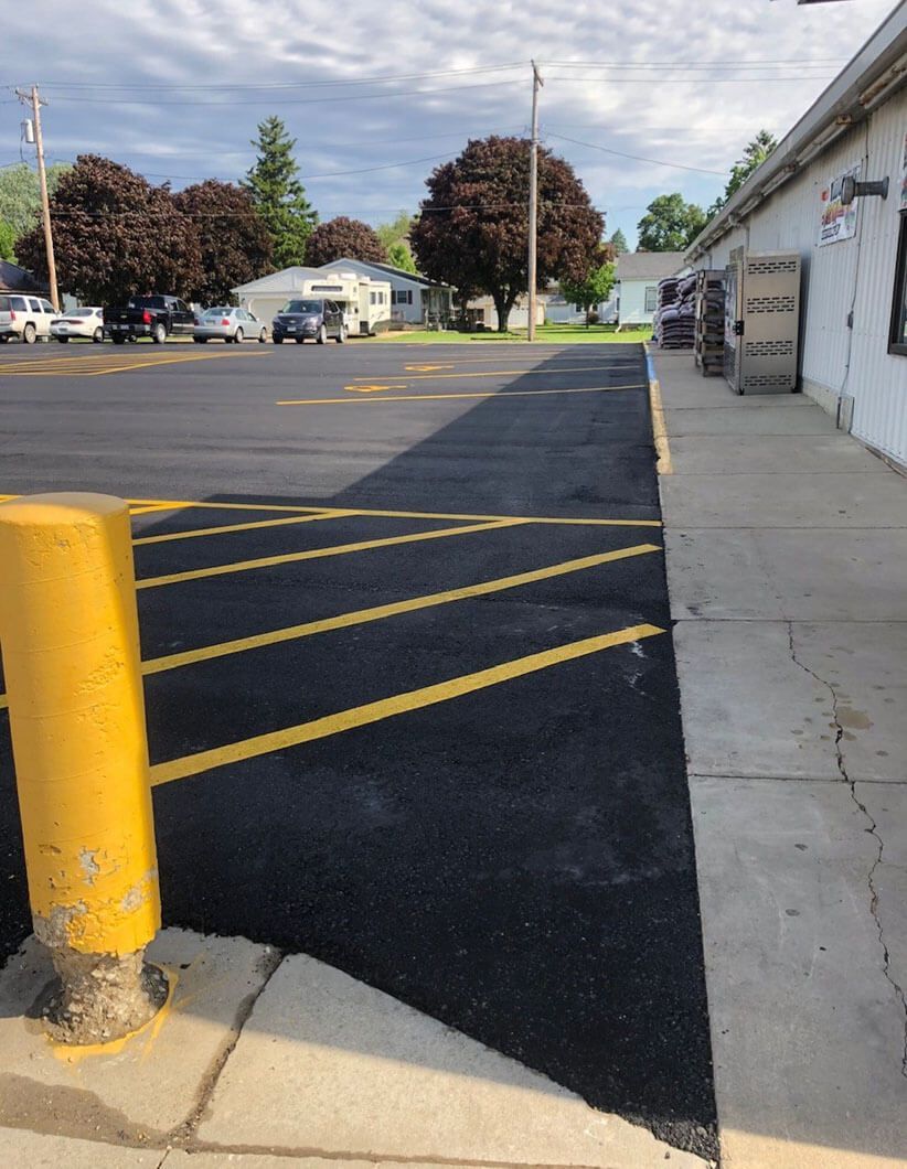 Newly paved asphalt in a parking lot next to a building sidewalk, featuring yellow-painted no-parking striped markings.