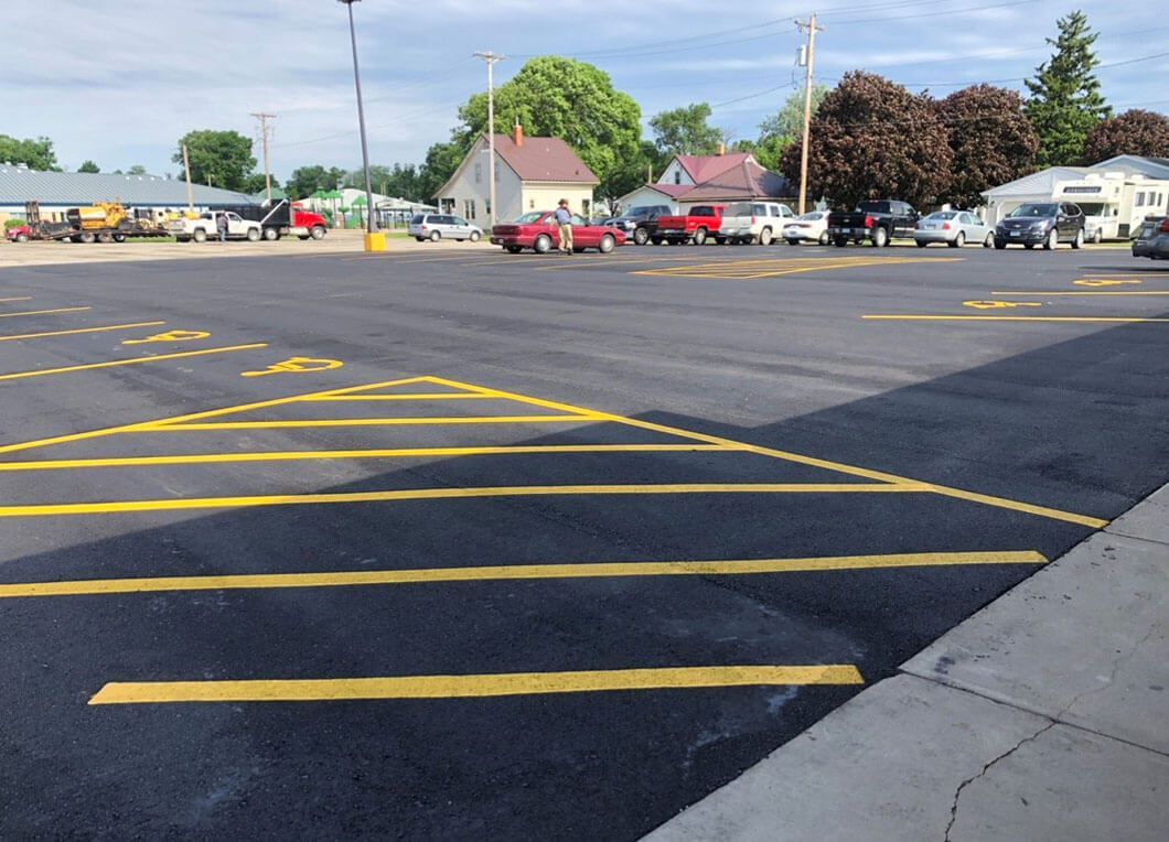 A paved parking lot with freshly painted yellow lines and hash marks, viewed from a sidewalk on a clear, sunny day.
