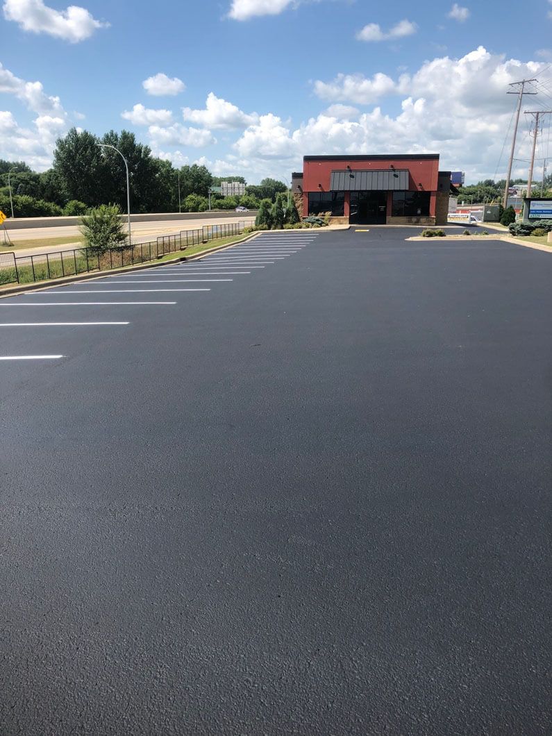 A freshly paved, black asphalt parking lot with white painted lines leading to a small commercial building under a blue sky.