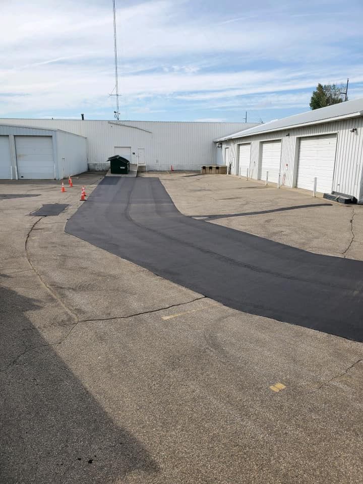 A freshly paved, black asphalt path cuts through a light gray concrete parking lot near several white warehouse doors.