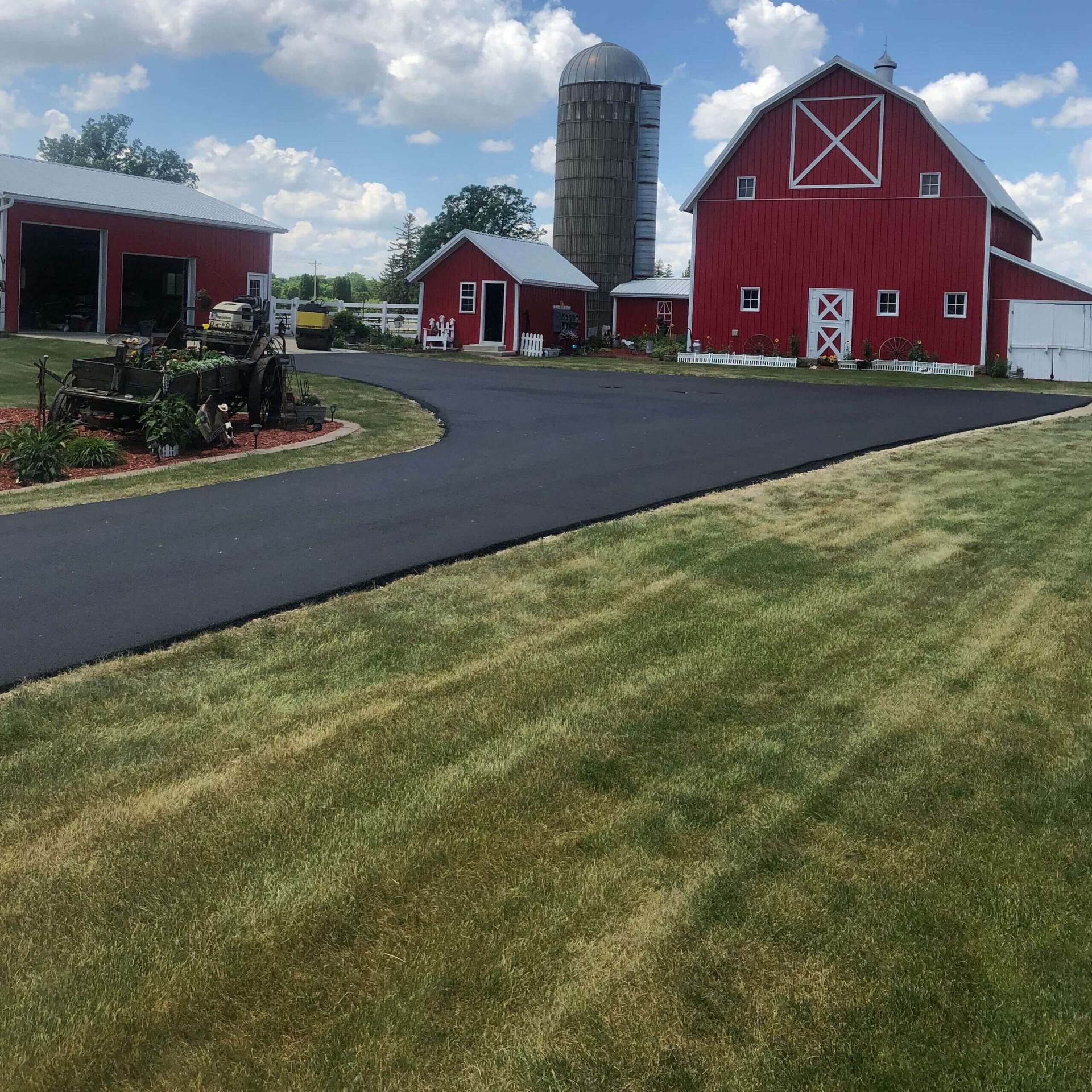 A black asphalt driveway leads toward a red barn and silo on a sunny, grassy farm property.