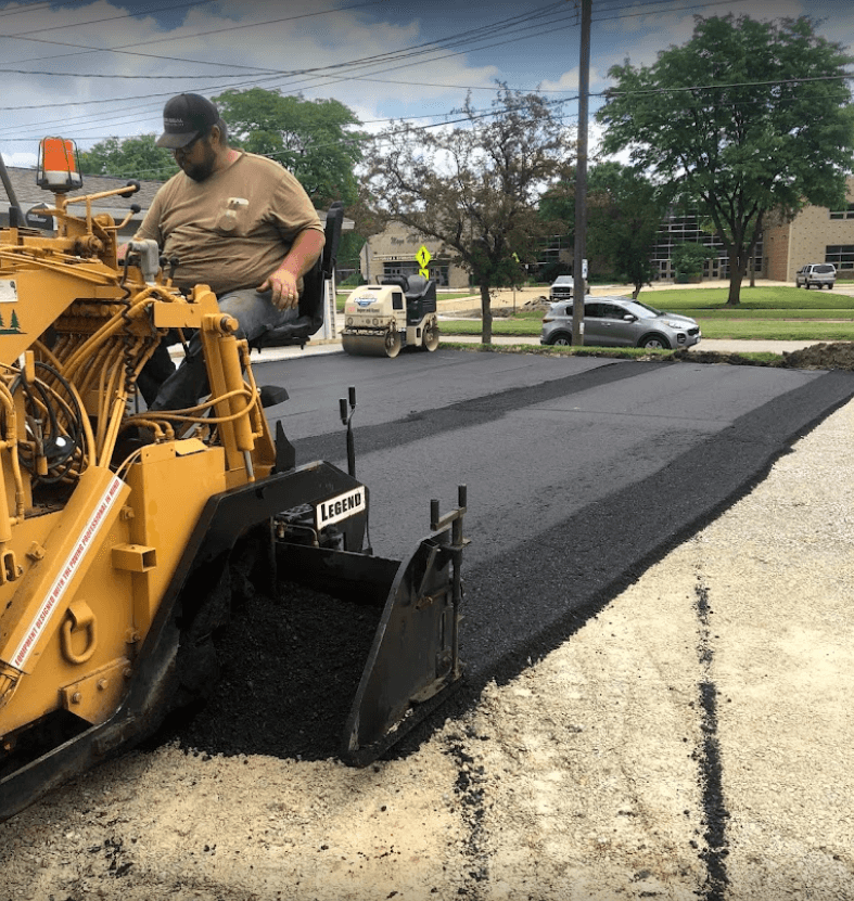 A worker operates a yellow asphalt paving machine, laying a fresh, smooth strip of black pavement on a sunny day.
