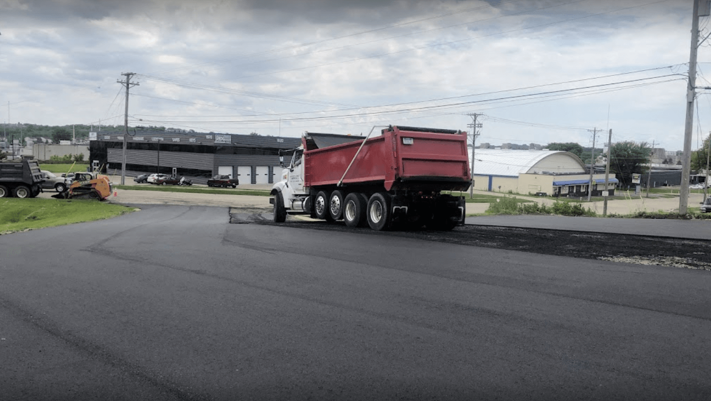 A red dump truck spreading fresh black asphalt on a newly paved parking lot under a cloudy sky.