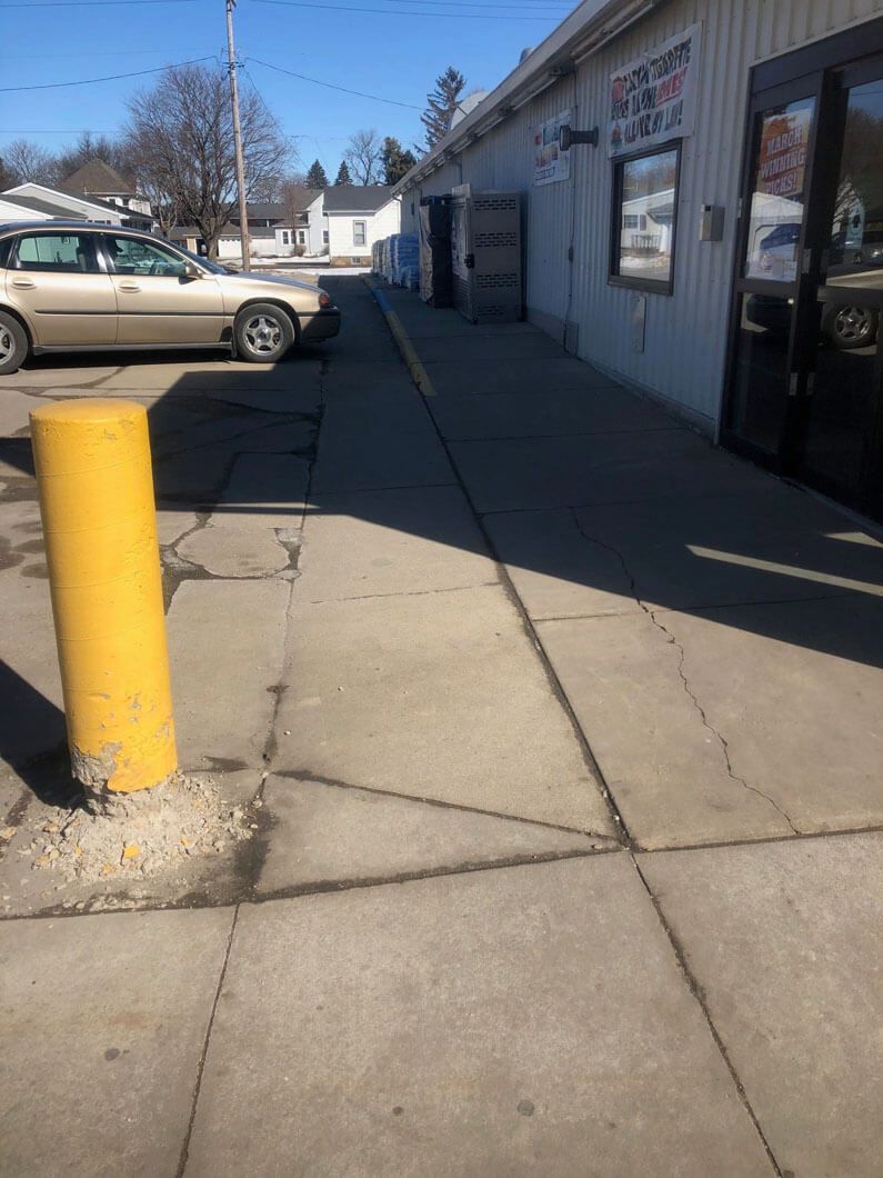 A sidewalk next to a commercial building with a yellow bollard in the foreground and a parked car on the left.