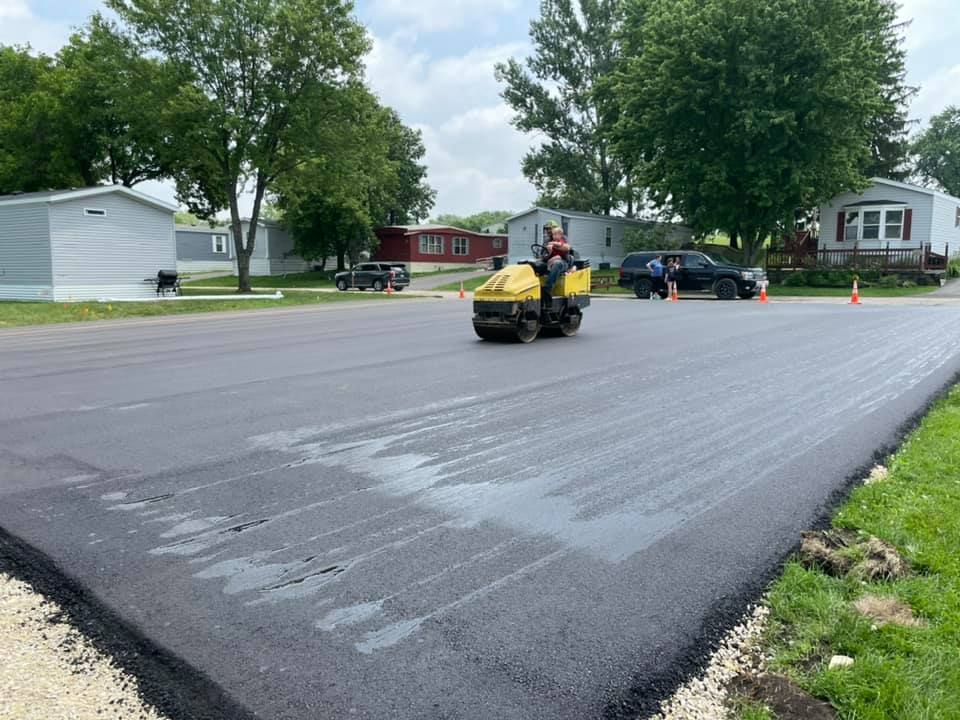 A yellow construction roller smooths freshly laid black asphalt on a road in a residential area.
