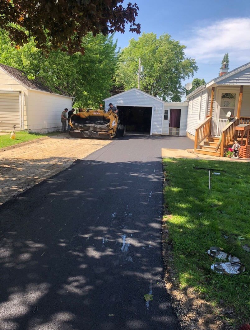 Workers use a construction vehicle to pave a fresh black asphalt driveway between two houses and a detached garage.