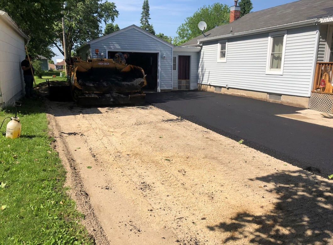 A half-paved driveway leads to a garage and house, with a sandy, unfinished section in the foreground.