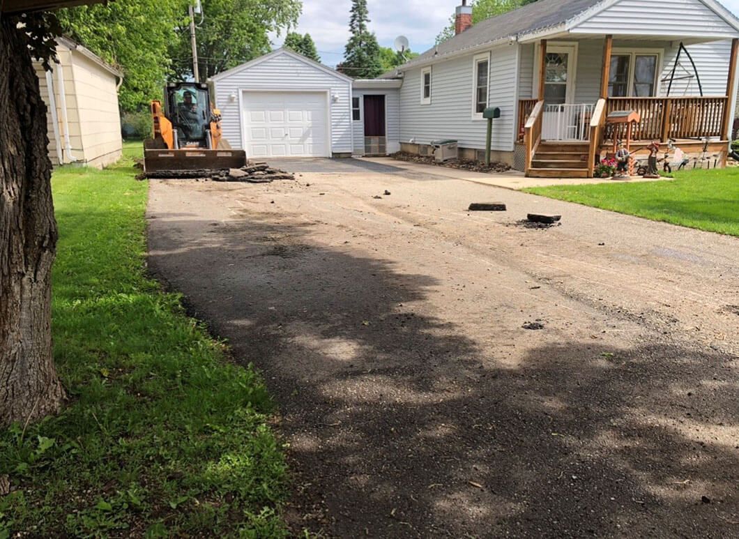 A skid steer loader sits on a driveway in front of a white house with a detached garage and a wooden porch.