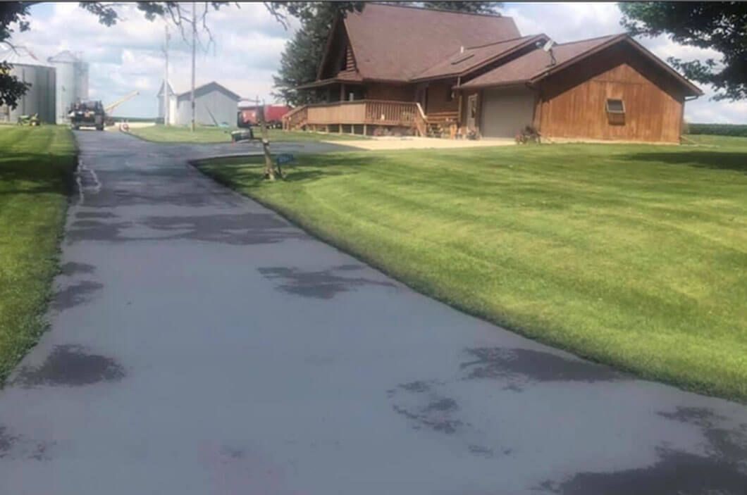 A paved driveway leads to a wooden house with a brown roof, surrounded by a large green lawn on a sunny day.