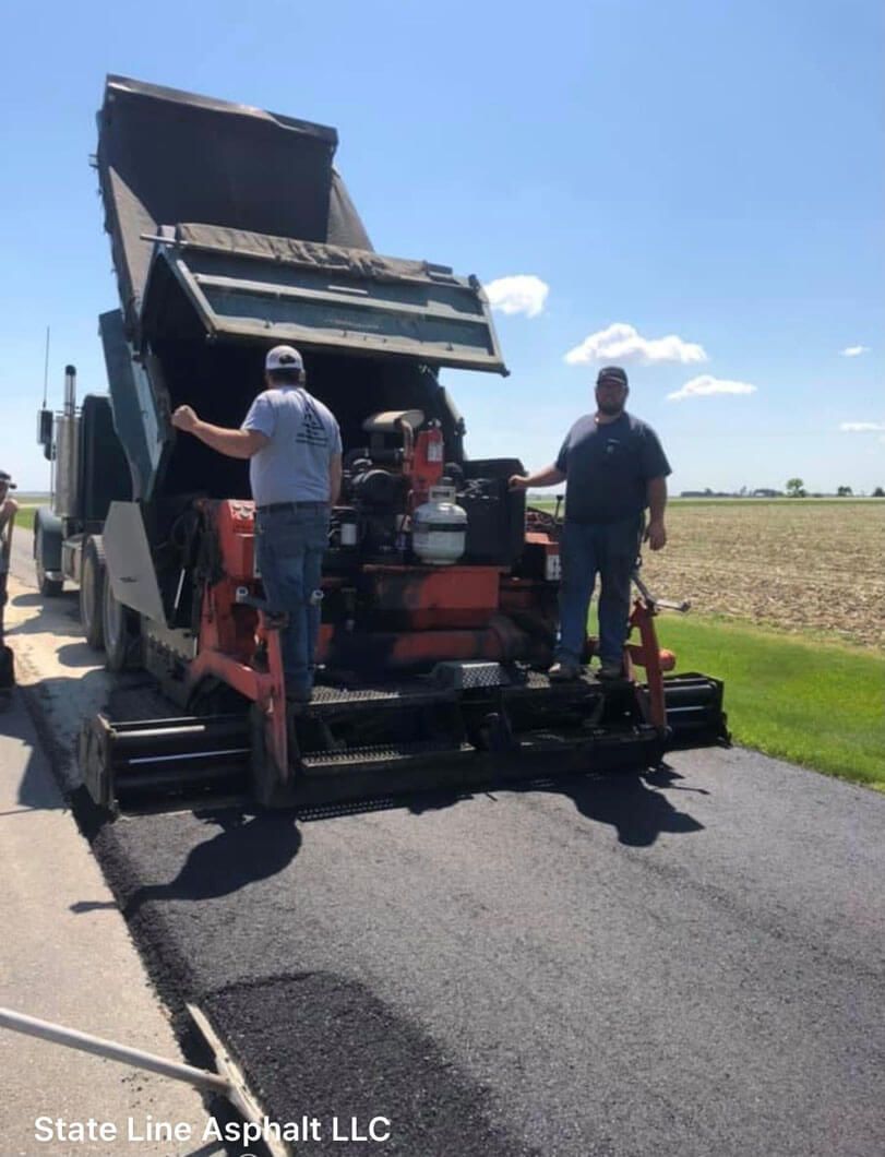 Two workers stand on an asphalt paving machine being filled by a dump truck on a rural road.