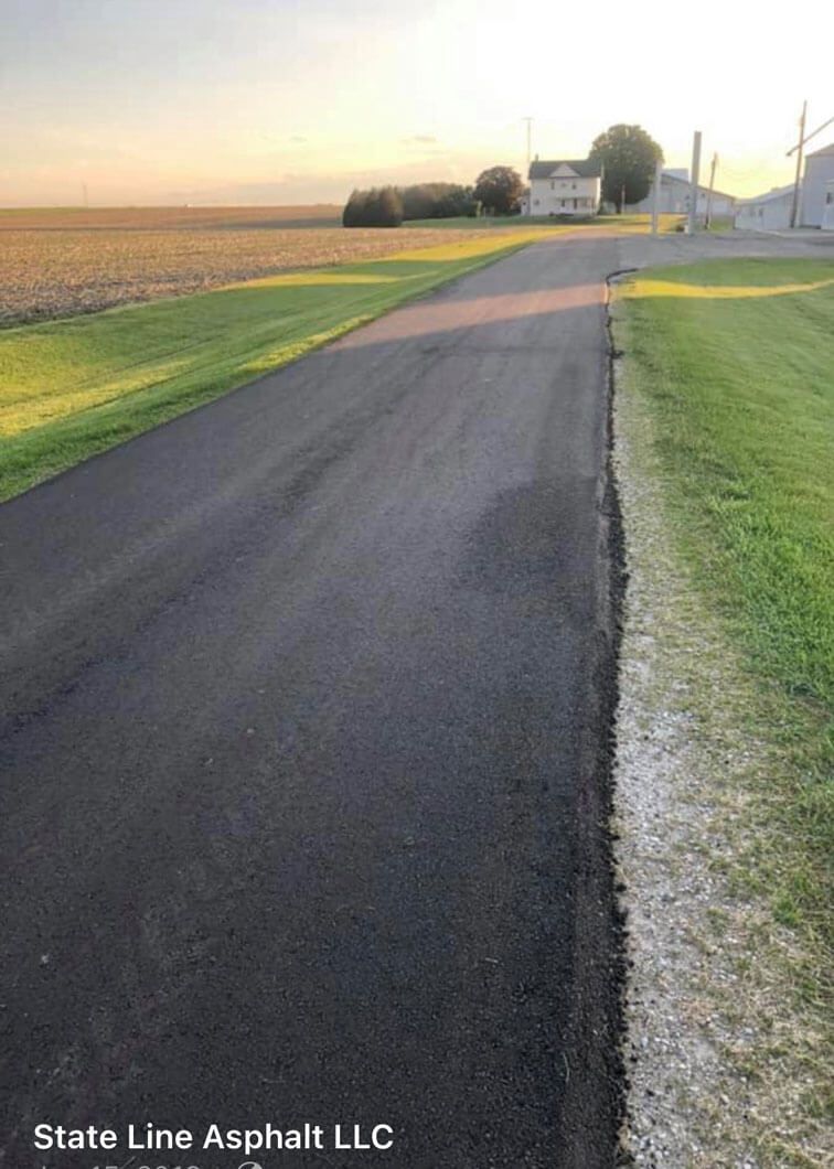 A newly paved asphalt driveway stretches toward a house in a rural setting at sunset.