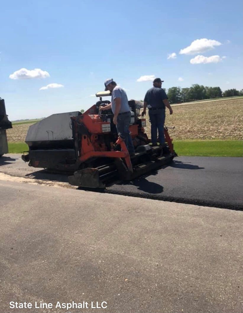 Two workers operate an orange asphalt paver on a road under a bright blue sky.