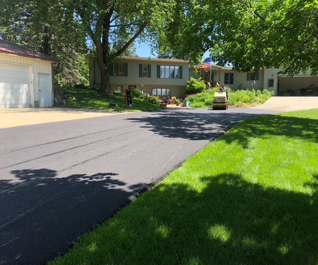 A newly paved asphalt driveway in front of a house, with a yellow steamroller parked near the front entrance.
