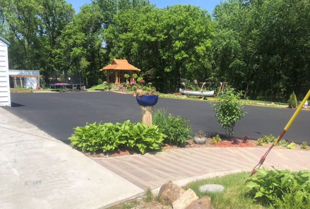 A freshly paved driveway leads to a wooden gazebo in a sunny, green backyard garden with landscaping and a small boat.