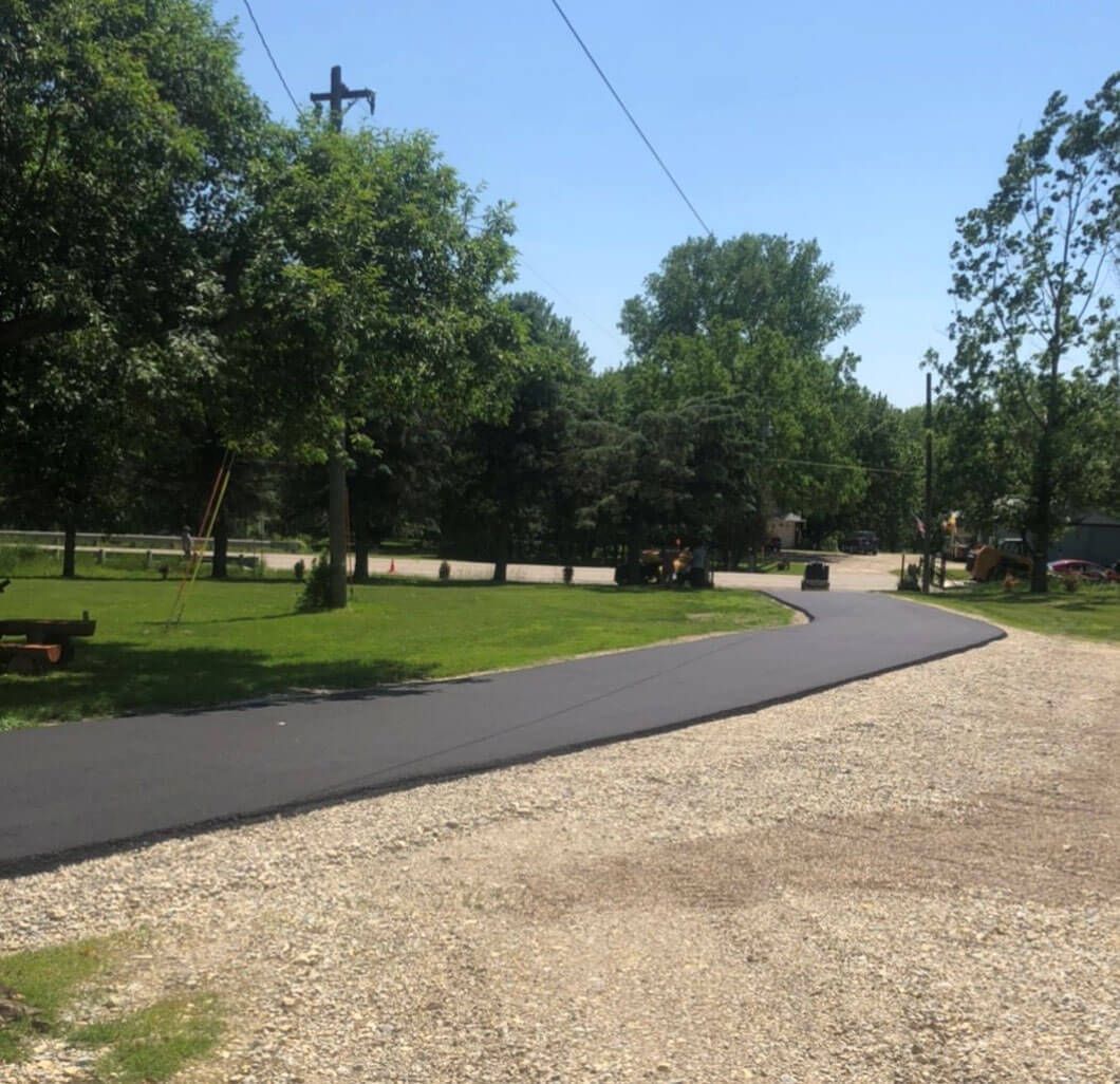 A freshly paved black asphalt driveway curves through a sunny, tree-lined residential area with a gravel foreground.