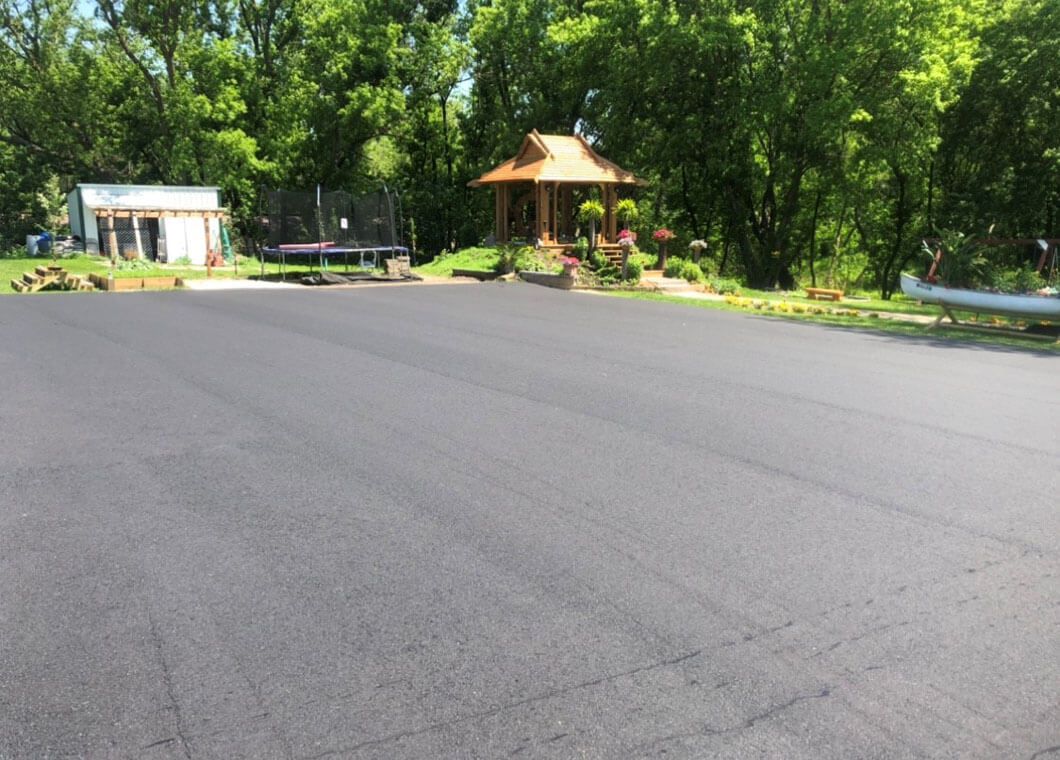 A freshly paved asphalt driveway leading toward a backyard with a gazebo, small shed, and trees.