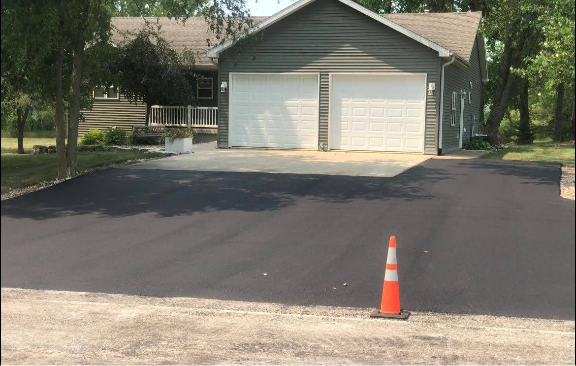A freshly paved dark asphalt driveway leads to a suburban house with a two-car garage, marked by an orange traffic cone.