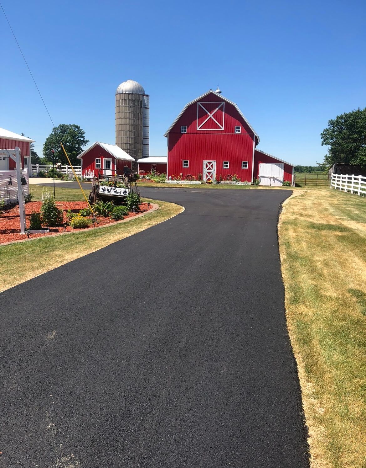 A paved driveway leads toward a red barn, a metal silo, and a small white building under a clear blue sky.