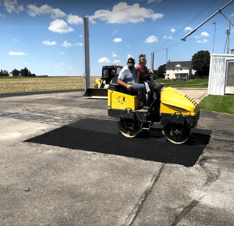 A person operating a yellow asphalt roller to compact a new black patch on a paved area near a farmhouse.