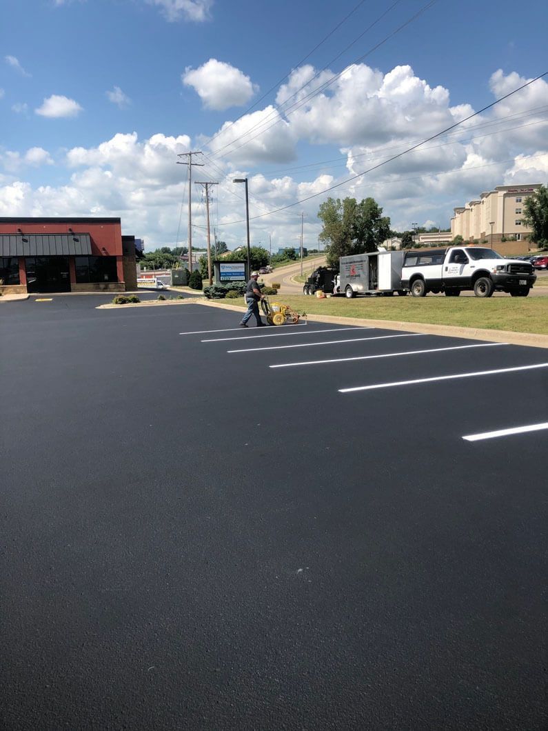 A person uses a line-striping machine to paint white parking space markers on a freshly paved black asphalt lot.