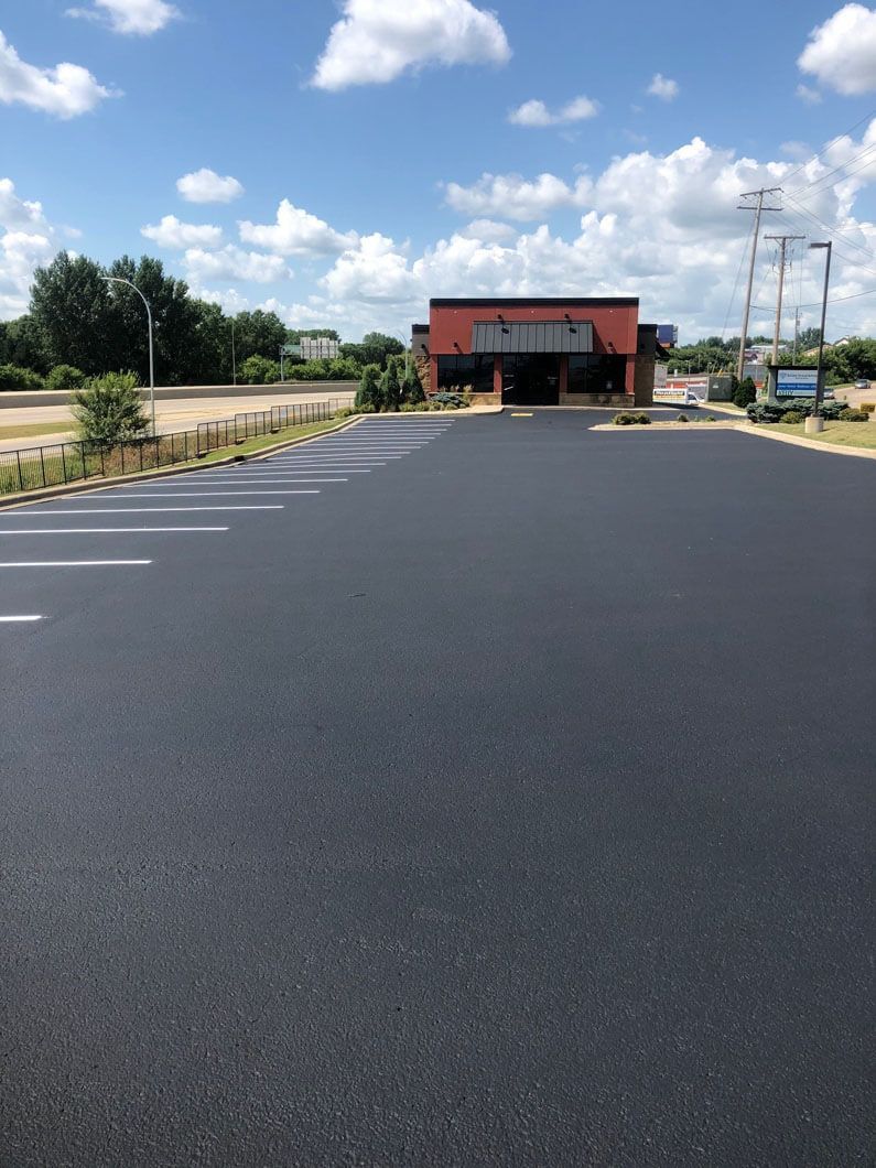 A freshly paved, empty asphalt parking lot with white painted stall lines leads to a low-profile brick commercial building.