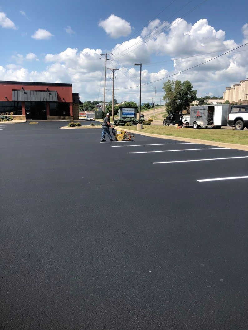 A person paints white parking stall lines on a newly paved asphalt lot next to a commercial building and trailer.