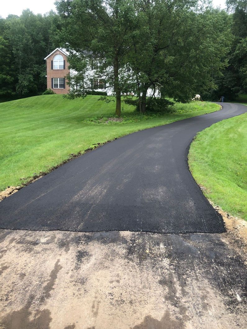 A freshly paved asphalt driveway curves through a green, grassy lawn toward a multi-story house surrounded by trees.