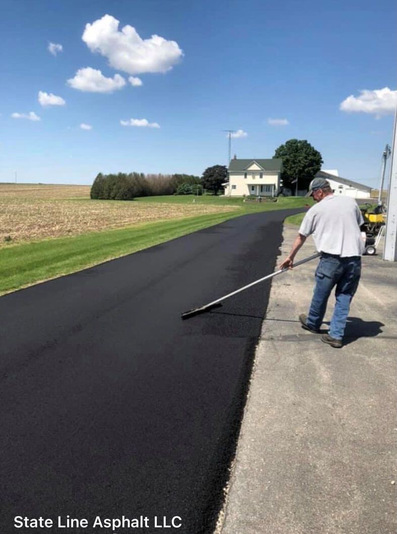 A worker uses a tool to spread fresh, black asphalt on a driveway beside a grassy field and a house under a blue sky.