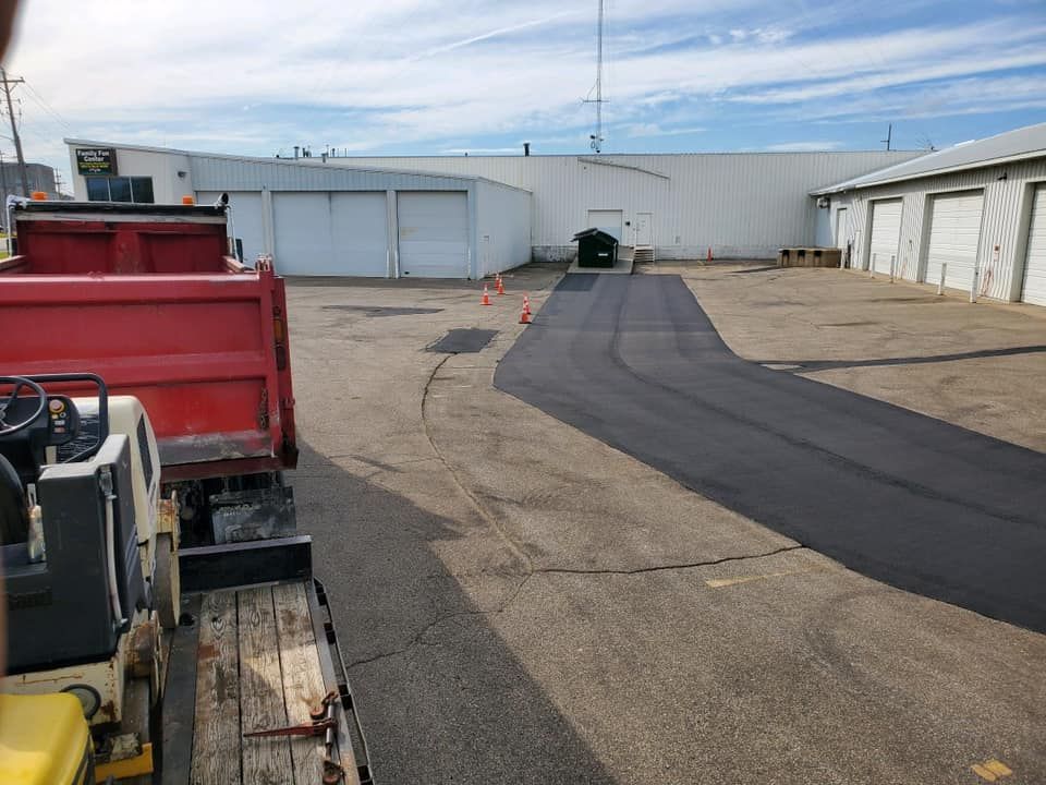 A red dump truck and construction equipment parked near a newly paved asphalt section in a commercial parking lot.