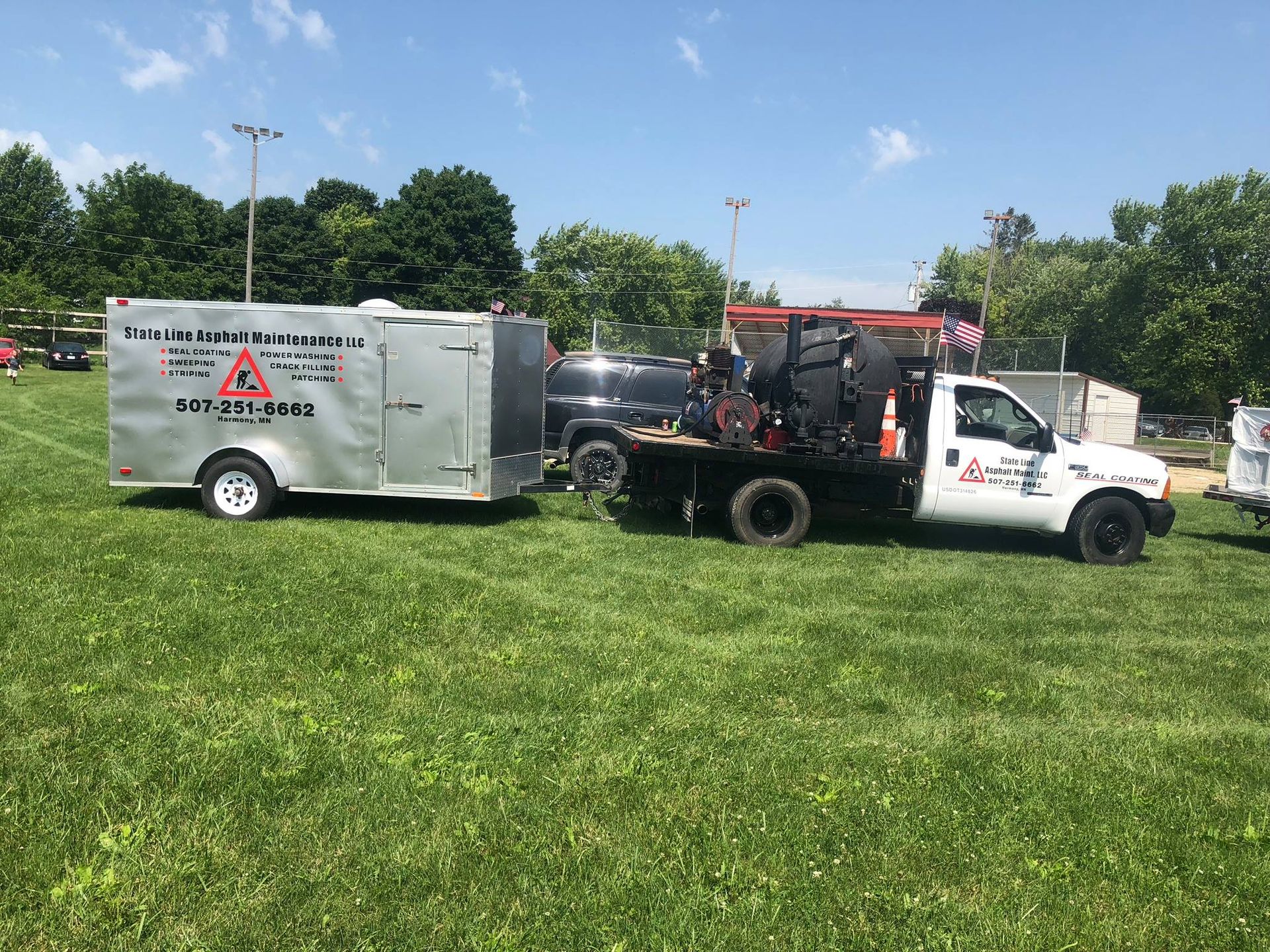 A white service truck towing a silver trailer parked on a grassy field under a sunny sky.