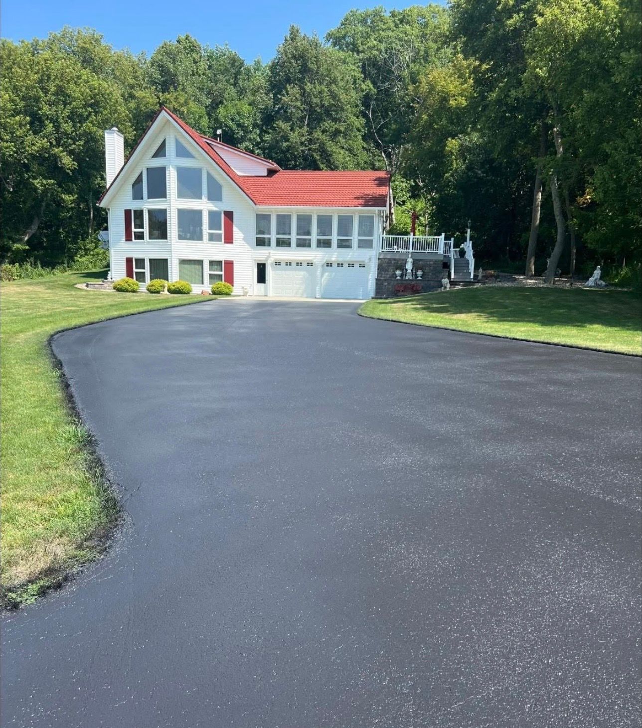 A worker spreads fresh black asphalt on a driveway in a rural setting under a clear blue sky.
