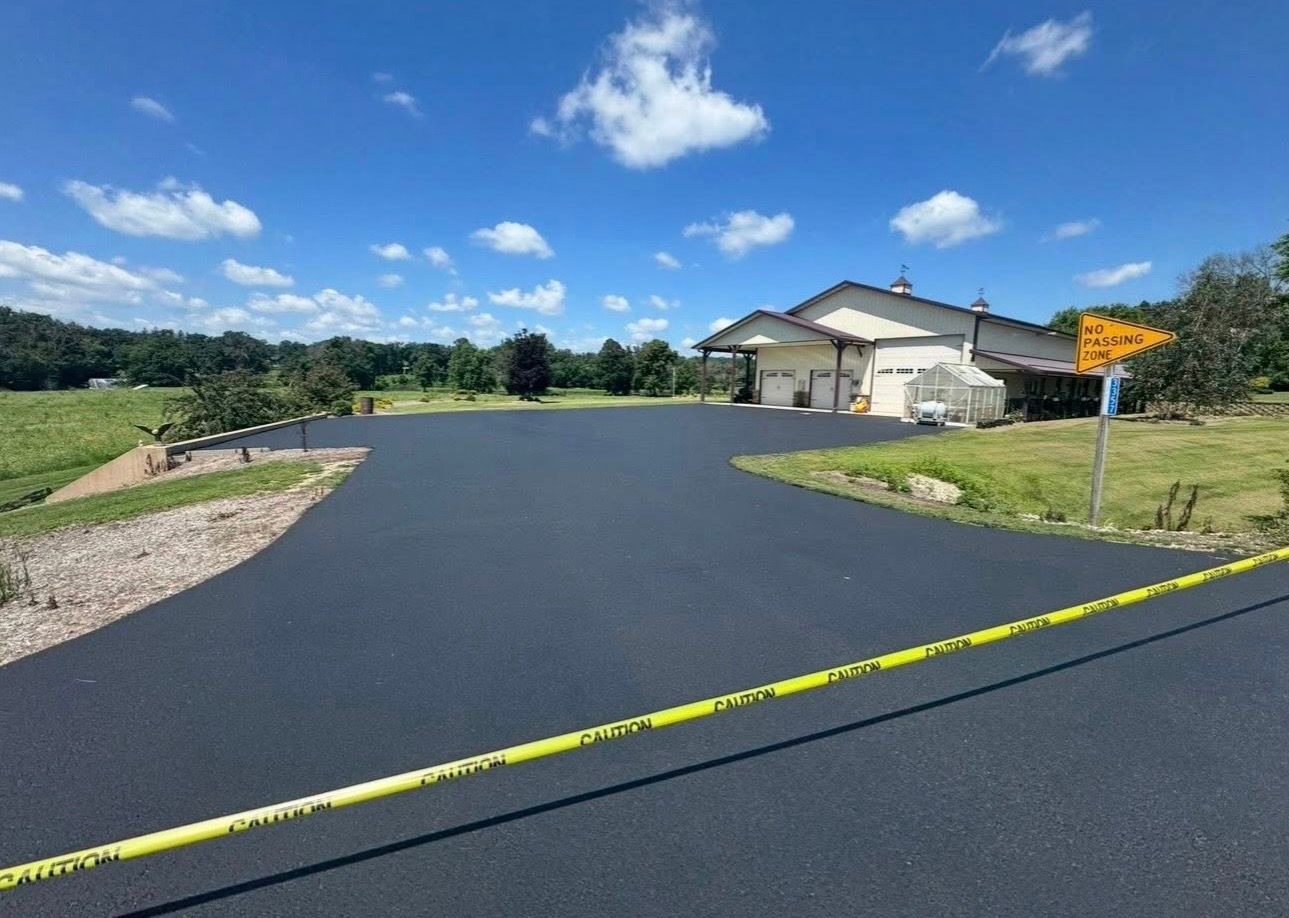 A modern white house with a red roof sits at the end of a freshly paved, smooth black asphalt driveway surrounded by trees.