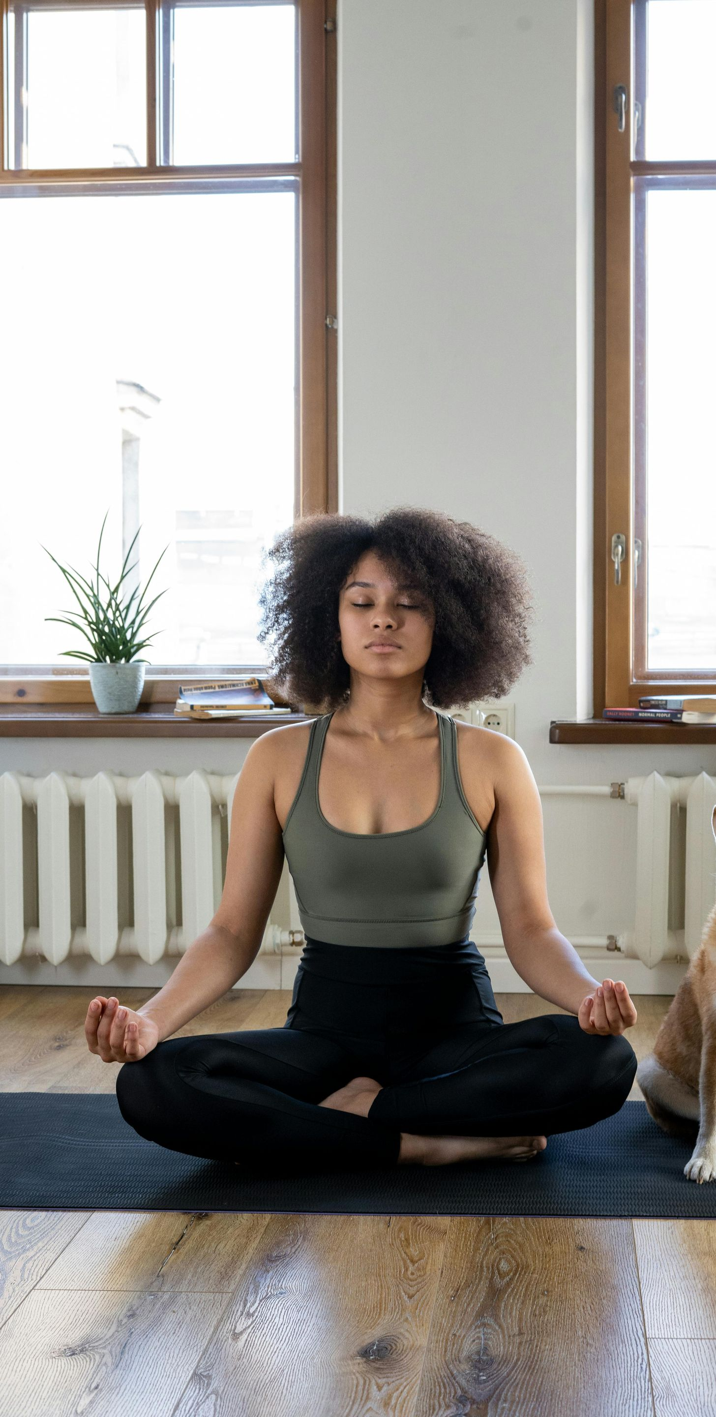 Vrouw in sportkleding zit met gekruiste benen, ogen gesloten, binnenshuis te mediteren.