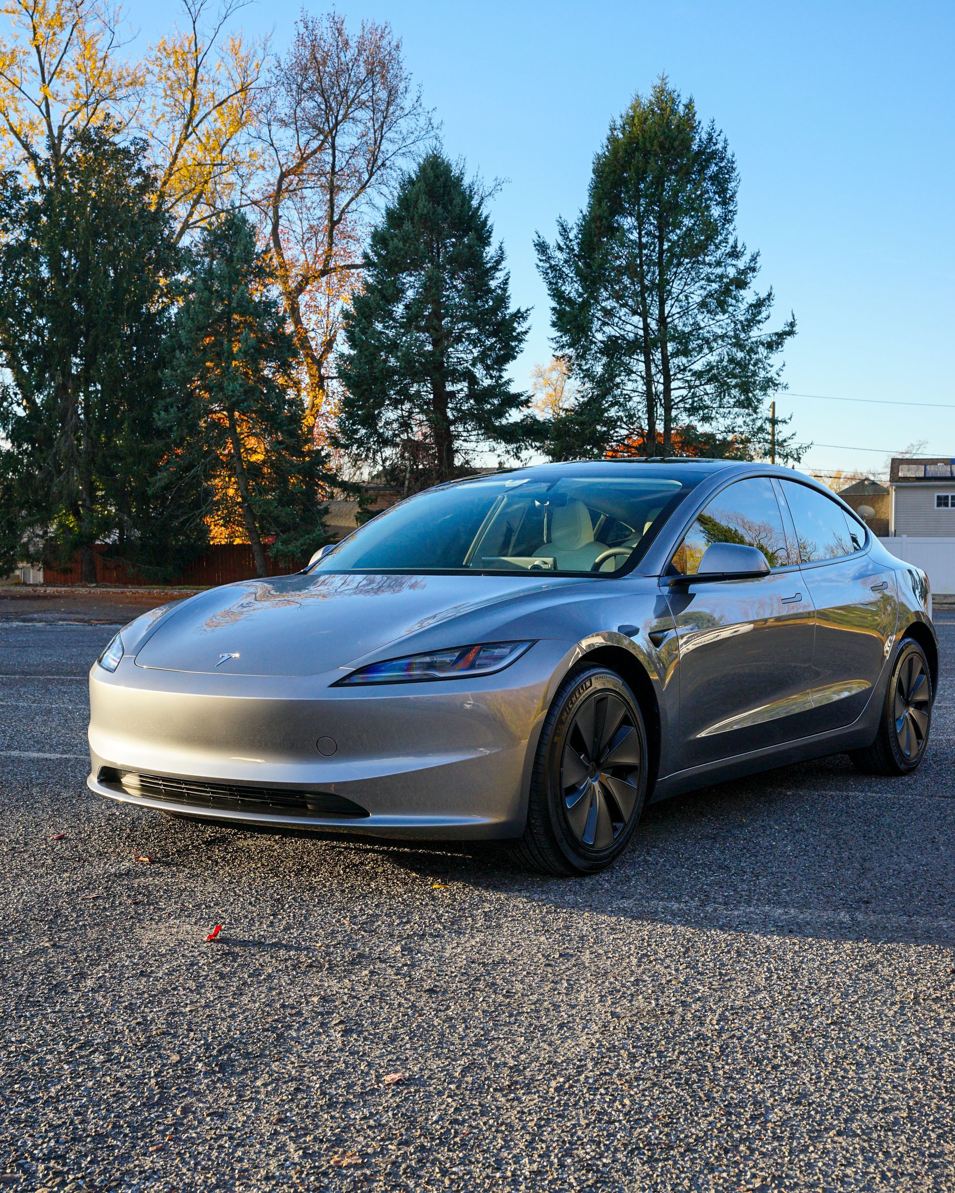 A silver tesla model 3 is parked in a gravel lot.