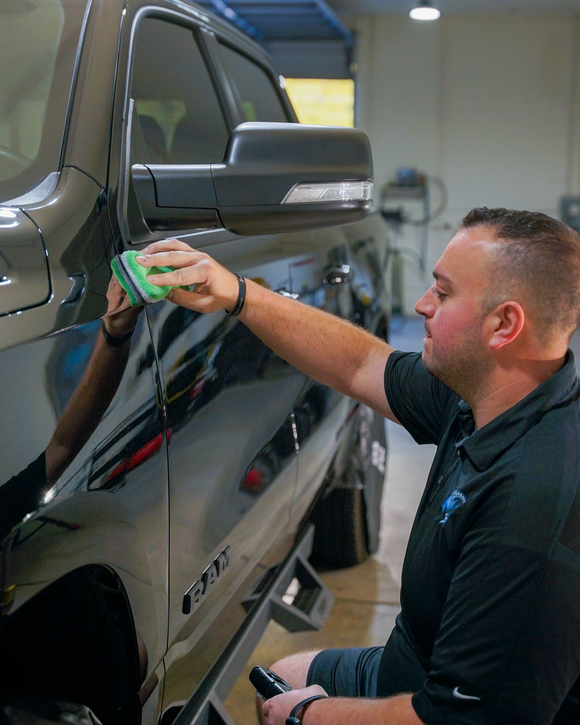 A person in a dark shirt applies a detailing product to the side of a black truck using a small, green foam sponge.