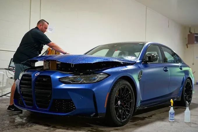 Technician installing PPF (paint protection film) on a blue BMW M3's front hood and bumper