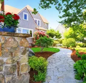 Stone pathway leads to a gray house with a lush garden, red flowers, and blue sky.
