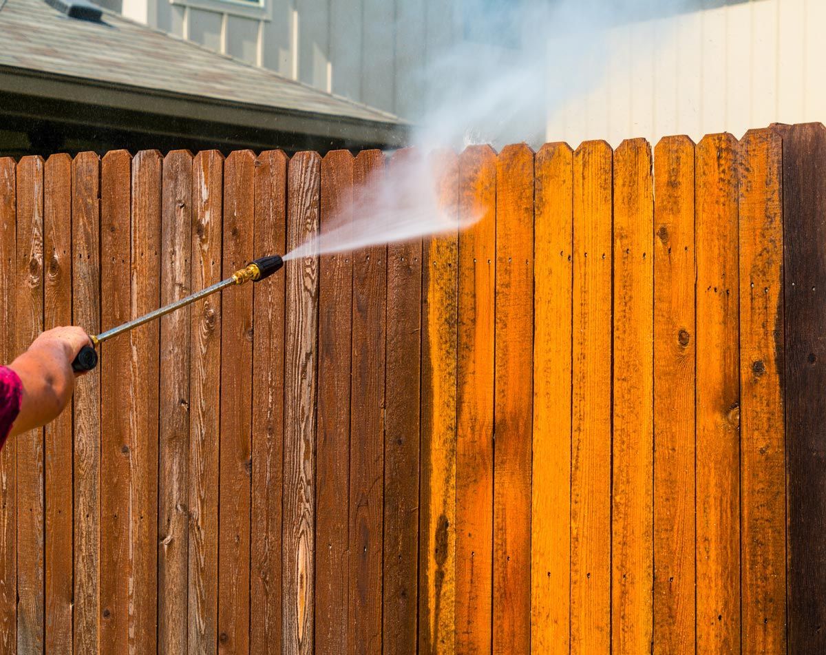 A person is using a high pressure washer to clean a wooden fence.