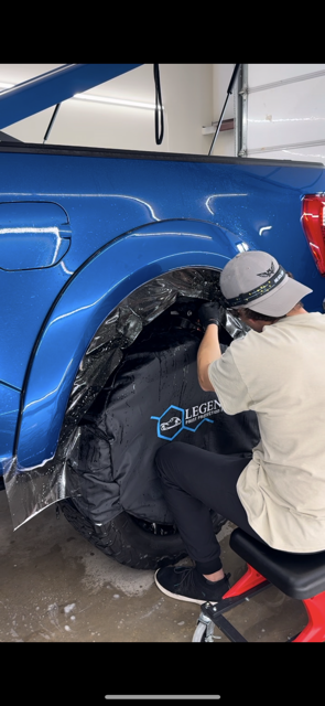 A person wearing a hat and working on a blue car's wheel well. They are applying a black protective layer.