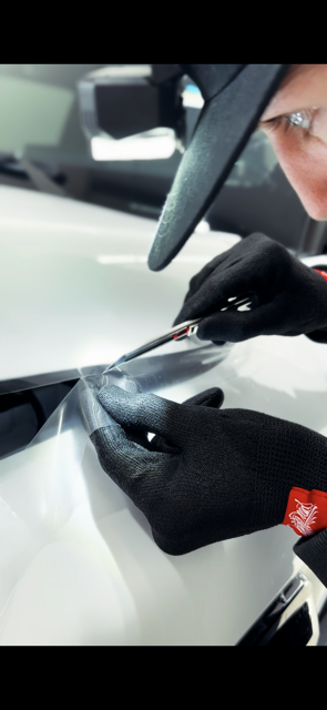 A person wearing black gloves uses a utility knife to trim film on a white car hood.